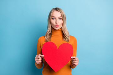 Photo of shiny flirty young lady wear orange sweater holding big heart sending you kiss isolated blue color background