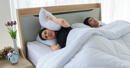 Asian young wife cannot sleep as her husband snoring beside her, woman closing ears with a pillow.