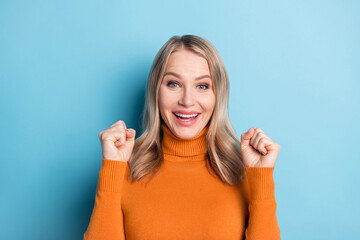 Photo of cute lucky young lady wear orange sweater smiling rising fists isolated blue color background