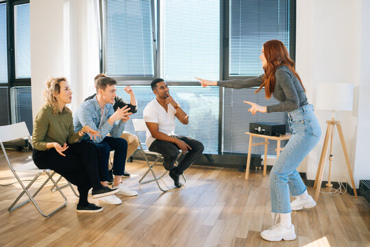 Side View Of Cheerful Redhead Woman Playing Charades With Friends Showing Pantomime By Window In Office. Group Of Activity Diverse Multi-ethnic Colleagues Playing In Active Games During Team Building.