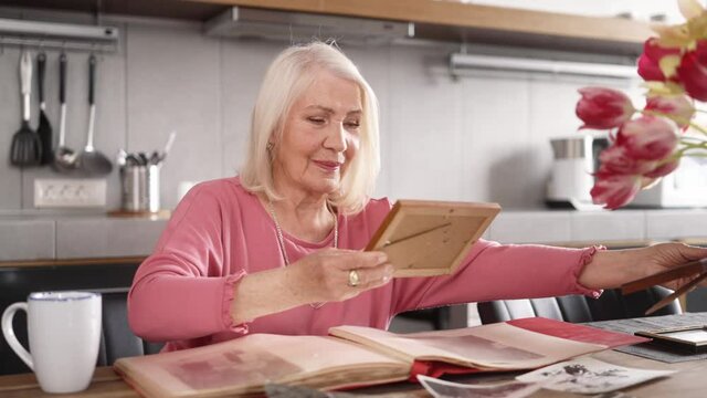 An elderly woman looking at framed photos over a cup of tea while sitting in the kitchen