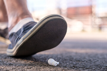 Foot of a man in shoes steps on a lost wireless headphones, which lies on the asphalt sidewalk,...