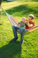 portrait of attractive nerd  man  with glasses in the park  with green lawn have a nice sunset with  a baby boy next to the hammock  . Happy fatherhood