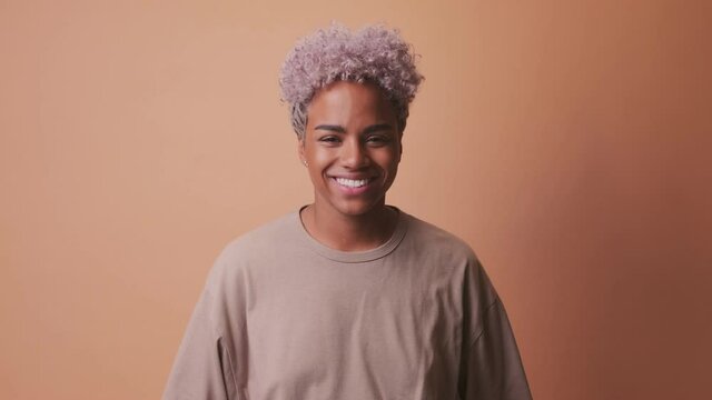 Close up view of curly haired young dark skinned woman holds hand to cheeks breath makes funny grimace plays fool isolated over brown background. Face expressions concept
