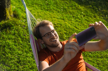 portrait of attractive nerd   man with glasses in the park with green lawn have a nice sunset in  the hammock  . Happy procrastination with phone