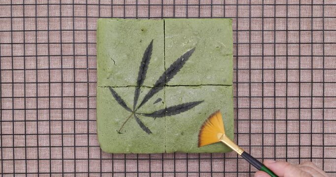 A cook using a brush to clean a surface of newly baked matcha cannabis white chocolate cake on a cooling rack.