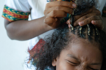 Close up African hairstylist braided hair of afro little girl, young girl makes a painful...