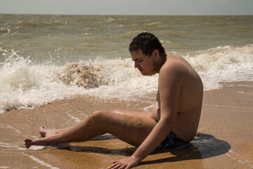 the guy is sitting on the seashore with his feet in the waves. the guy with autism