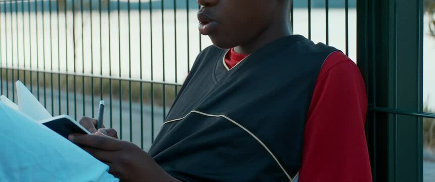 Black African American teenager boy writing lyrics for his song on a basketball court. Aspiring rap artist dreaming of becoming future star. Shot with 2x anamorphic lens