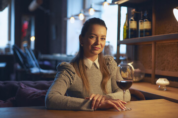 A beautiful young woman sits at a wooden table in the premises of a wine bar and drinks wine from a...
