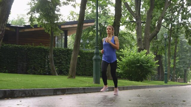 Slow motion woman in sportswear running in park on wet walkway. Tracking shot fit female training outside after rain. Concept of fitness