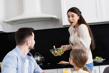 amazed woman touching chest while holding vegetable salad near blurred family