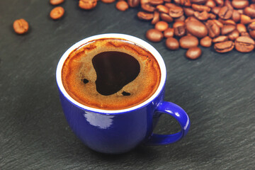A blue cup of coffee on a black table next to fresh roasted coffee beans