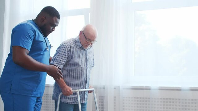 African-American caregiver is teaching disabled old man to walk with walker. Professional nurse and handicapped patient in a nursing home. Assistance, rehabilitation and health care.