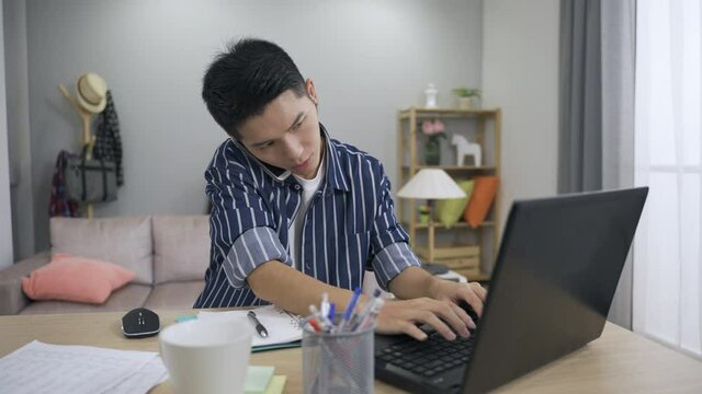 Asian Male Worker Is Holding The Phone Between His Ear And Shoulder To Look Up Information On The Computer And Taking Notes With A Pen While Working From Home.