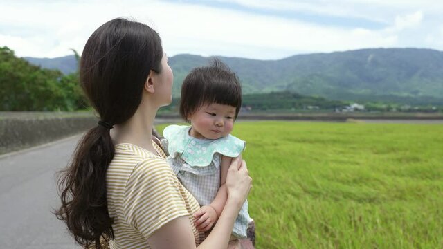 side portrait mother is gently stopping her baby from putting hand into mouth while enjoying the peaceful landscape near a pastureland on a breezy sunny day.
