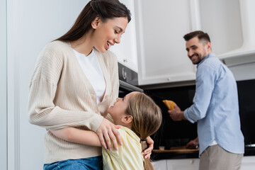 Fototapeta premium excited woman embracing daughter near blurred husband holding bread in kitchen