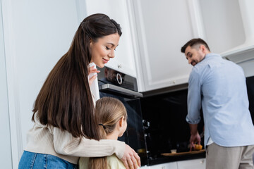 happy mother hugging child near blurred husband cutting bread in kitchen