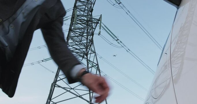 Businessman takes charging cable off electromobile near tower with power transmission lines under blue sky closeup slow motion