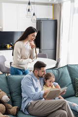 woman standing near husband and daughter reading book on sofa at home