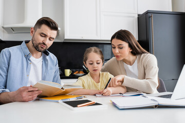 man reading book and woman pointing with finger while helping daughter doing homework