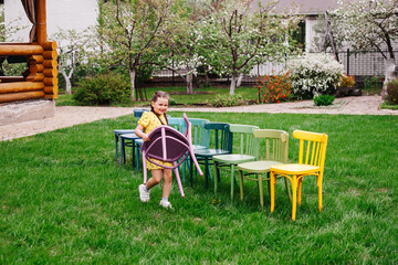 a girl in a yellow dress carries a wooden chair and arranges chairs in a line, organizing a children's birthday party outdoors in the garden in summer.  - obrazy, fototapety, plakaty