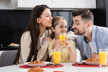 Obraz na płótnie Canvas joyful parents embracing daughter during breakfast in kitchen