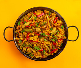 Chicken paella on yellow background, a traditional Valencian (Spanish) dish made of rice with chicken and vegetables in a pan. view from above, flat lay