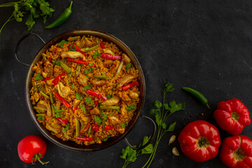 A traditional Chicken paella on dark background with tomatoes and parsley, made of rice with chicken and vegetables in a pan. view from above, flat lay. Copy space