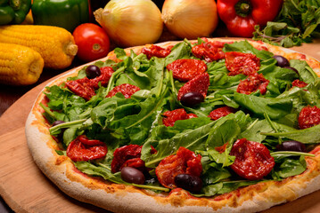 Arugula and sun dried tomato pizza on wooden board and vegetables in the background.