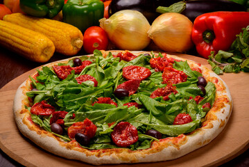 Arugula and sun-dried tomato pizza on wooden board and vegetables in the background.