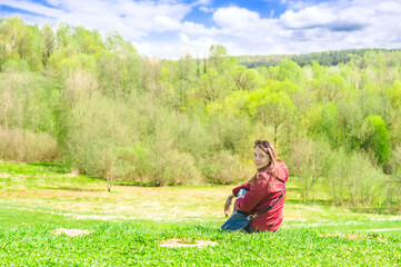 Naklejka na ściany i meble Smiling woman in red coat sitting outdoors on the grass in front of forest landcape and looking at camera