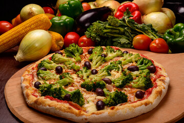 Broccoli pizza with black olives on wooden board and vegetables in the background.