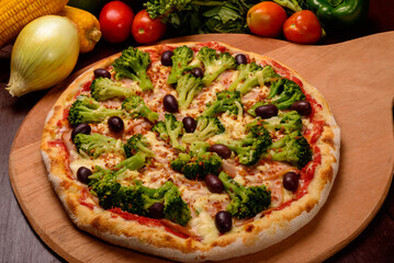 Broccoli pizza with black olives on wooden board and vegetables in the background.