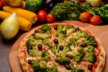 Broccoli pizza with black olives on wooden board and vegetables in the background.
