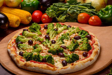 Broccoli pizza with black olives on wooden board and vegetables in the background.
