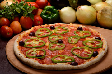 Pepperoni pizza with green peppers on wooden board and vegetables in the background.