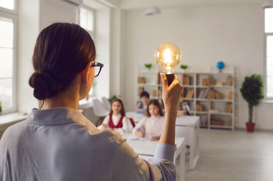 Student creativity thinking development, modern school education. View from back on teacher with idea light bulb inspires elementary children at lesson standing front of students on blurred background