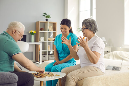 Happy Senior Patients And Home Care Nurse Play Checkers. Old Retired Couple And Caretaker Enjoy Intellectual Table Game To Keep Brain Young And Healthy. Leisure Activity As Disease Prevention Therapy