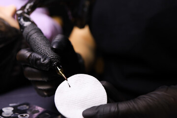hands of a tattoo artist wearing black gloves holding a tattoo machine in one hand and a cotton...