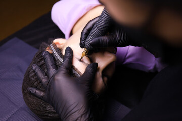 a tattoo artist wearing black gloves performs permanent eyebrow makeup. one hand holds the client's head the other works with a tattoo machine - obrazy, fototapety, plakaty