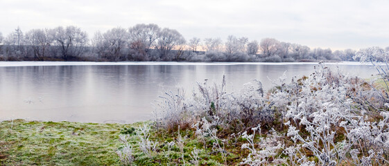 Winter landscape with frost-covered plants by the river and the reflection of trees in the water. Winter day, panorama