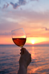 A glass of red wine in the hands of a girl in the rays of the sunset on the seashore. Alcoholic beverage tasting. Beautiful sunset with wine on the beach. Romantic evening aperitif. Glass closeup