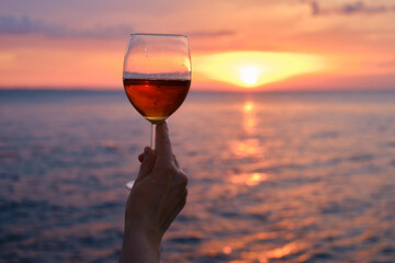 A glass of red wine in the hands of a girl in the rays of the sunset on the seashore. Alcoholic beverage tasting. Beautiful sunset with wine on the beach. Romantic evening aperitif. Glass closeup