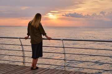 Beautiful girl stands by the sea and looks at the dawn. Girl in travel. Amazing scenic outdoors view. Beautiful dawn on the seashore. Adventure lifestyle