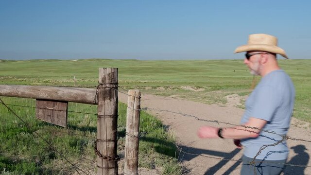 Senior man in a cowboy hat, a farmer or rancher, is opening barbed wire cattle gate on a ranch road, summer scenery with a green prairie in Colorado