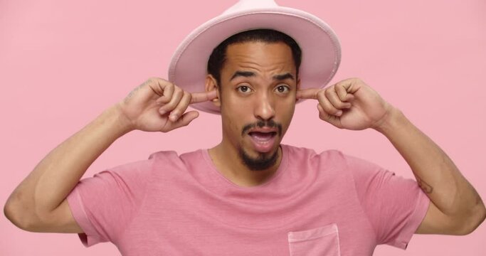 Displeased bearded black man covering his ears with fingers and talking to camera isolated over pink