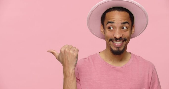 Happy black man in pink hat pointing at copy space and smiling isolated over pink