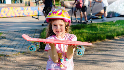 Sportive girl holding a pennyboard in her hands and smiling. Schoolgirl girl spends summer holidays in the park of extreme sports. Sport in children's life concept. Healthy lifestyle concept.