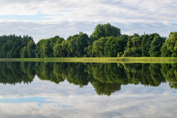Fototapeta na wymiar Lake shore in summer. Reflection of green trees and clouds in the water